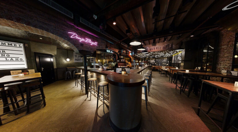 Interieur van Café van Bommel in Maastricht, met een sfeervolle bar, houten vloeren, industriële accenten en neonverlichting. De ruimte is ingericht met hoge tafels, krukken en een opvallende muurschildering aan de achterwand.