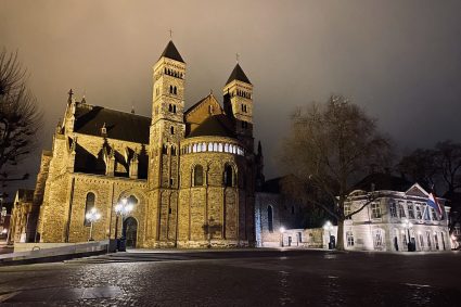 Sint Servaas Basiliek Een foto genomen in de avond van de Sint Servaas Basiliek in Maastricht. De basiliek, verlicht door zachte lampen, straalt een warme, gouden gloed uit tegen de donkere nachtelijke hemel. De karakteristieke Romaanse architectuur, inclusief de dubbele torens en ronde bogen, is duidelijk zichtbaar. Rechts op de foto staat een klassiek gebouw met de Nederlandse vlag trots wapperend in de wind. De lege straat voor de basiliek is geplaveid met kasseien, en de omgeving is rustig en verlaten, wat een serene en historische sfeer creëert.